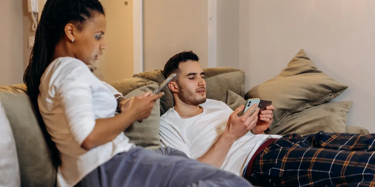 A couple relaxing on a sofa at home, one holding a phone and a plain payment card while the other looks at a tablet